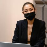 Professional woman in formal attire wearing a face mask, seated at a desk with a laptop.